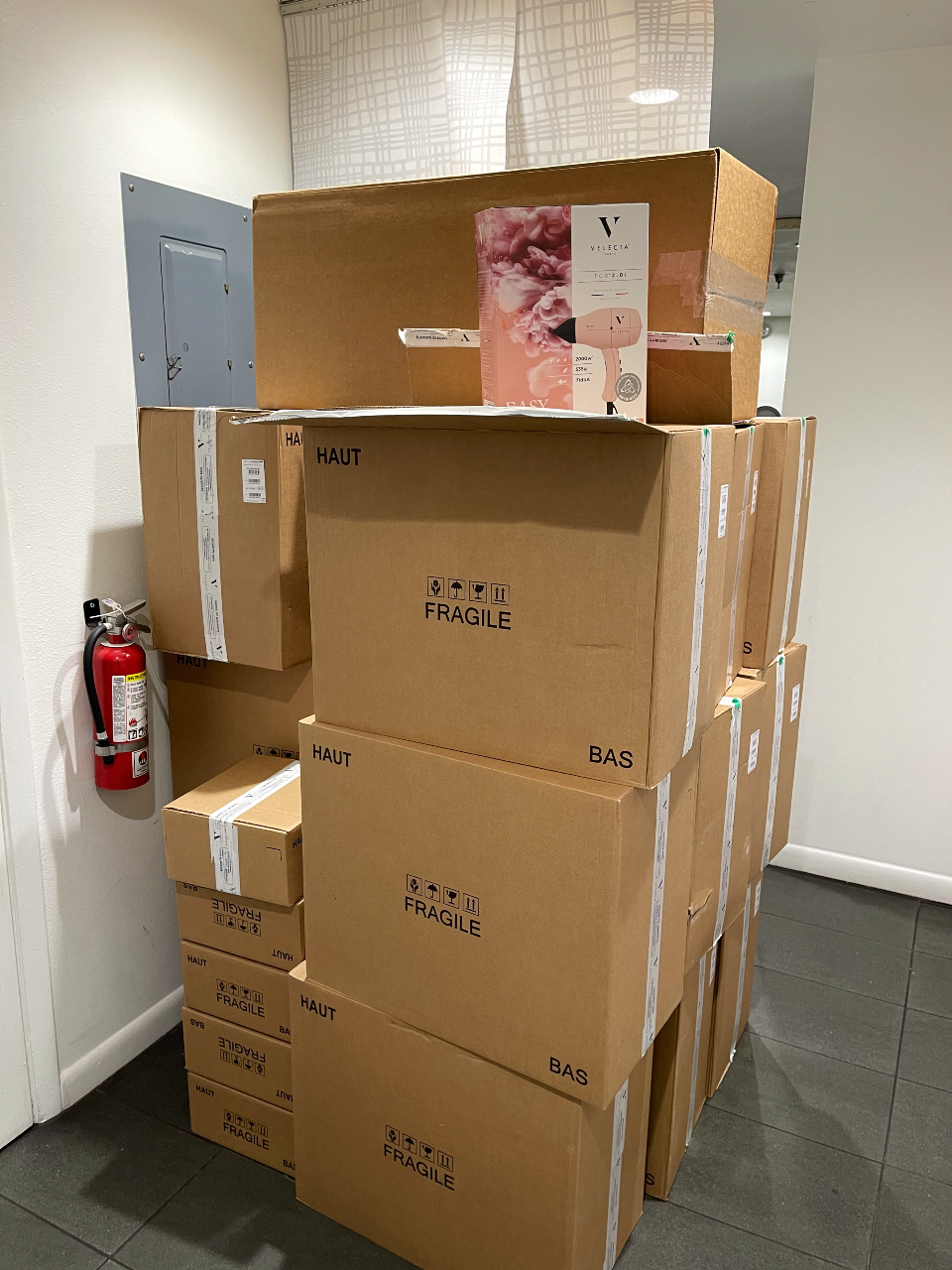 A stack of various-sized brown cardboard boxes labeled "FRAGILE" is piled against a wall next to a mounted fire extinguisher in a hallway. A perfume advertisement from Paris is placed on top of the boxes. - Nuovo Salon Group in Sarasota & Bradenton, FL