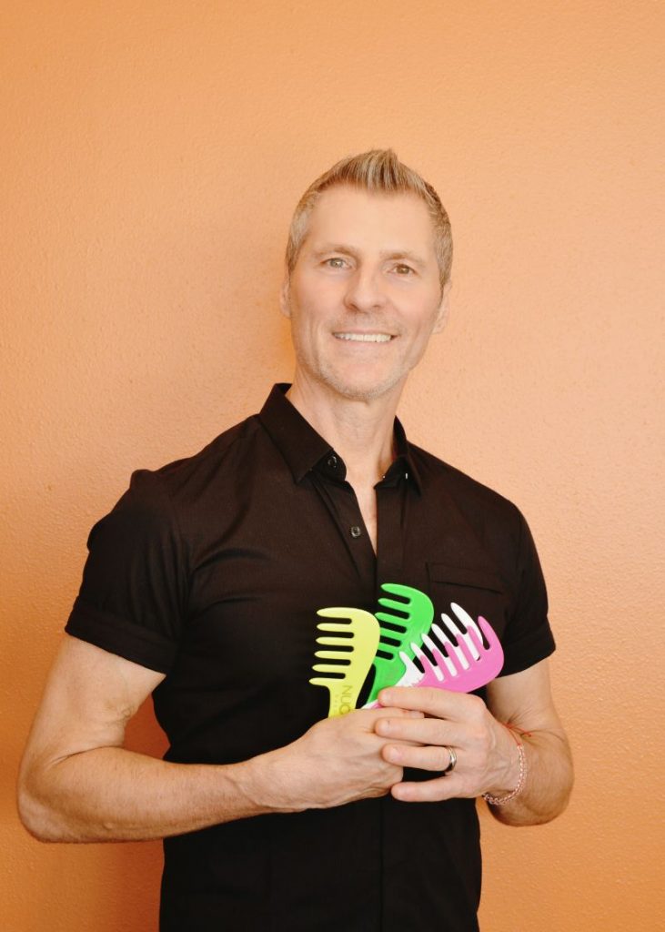 A man in a black shirt stands against an orange wall holding three colorful plastic combs, smiling as if he's ready to perfect his hairstyle. - Nuovo Salon Group in Sarasota & Bradenton, FL