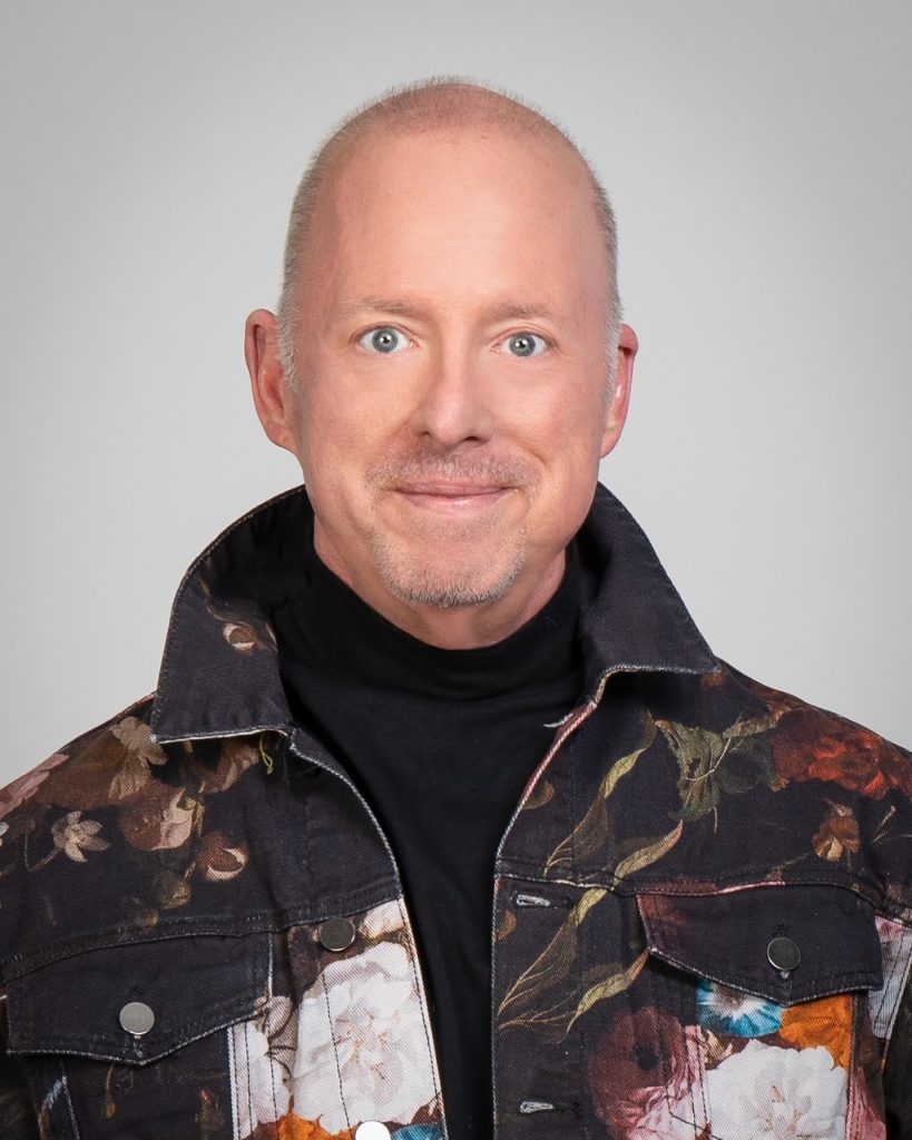 A man with a bald head and light facial hair wearing a black turtleneck and a jacket with a floral pattern stands against a plain gray background, showcasing his distinctive hairstyle. - Nuovo Salon Group in Sarasota & Bradenton, FL