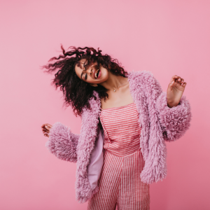 A person with curly hair, wearing a fluffy pink jacket and striped clothing, smiles and dances against a vibrant pink background, embodying bold colors and ahead of the curve fashion. - Nuovo Salon Group in Sarasota & Bradenton, FL