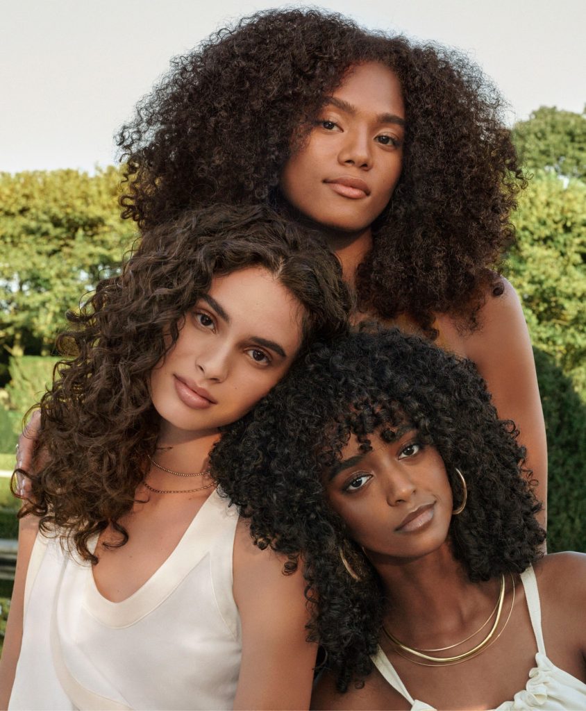 Three women with curly hair, wearing sleeveless tops and subtle jewelry, pose closely outdoors with greenery in the background. - Nuovo Salon Group in Sarasota & Bradenton, FL