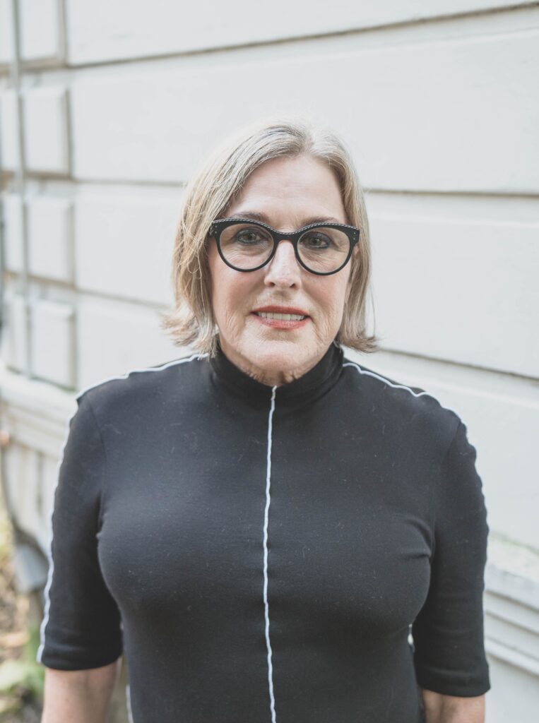 In Episode 1 of Opposites Align, a person with short hair and glasses stands outside in front of a white wall, sporting a stylish black top. - Nuovo Salon Group in Sarasota & Bradenton, FL