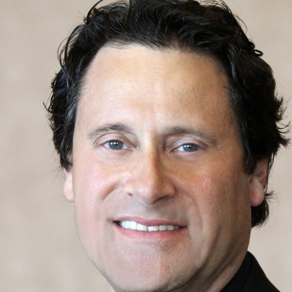 A man with short, dark wavy hair and light skin is smiling slightly while looking at the camera, as if inviting you to tune in. He is wearing a dark-colored shirt or jacket. - Nuovo Salon Group in Sarasota & Bradenton, FL
