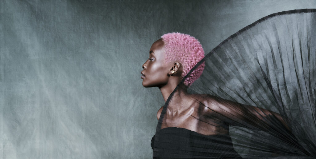 A person with short pink hair and gold earrings poses in profile against a gray background, wearing a NUOVO black outfit with sheer, pleated fabric—a striking look perfect for any holiday gift guide or self-care inspiration. - Nuovo Salon Group in Sarasota & Bradenton, FL