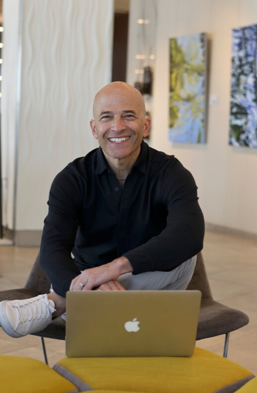 A smiling man sits cross-legged on a chair with a laptop on a small table in front of him, as opposites align in a modern indoor setting adorned with abstract art on the walls. - Nuovo Salon Group in Sarasota & Bradenton, FL
