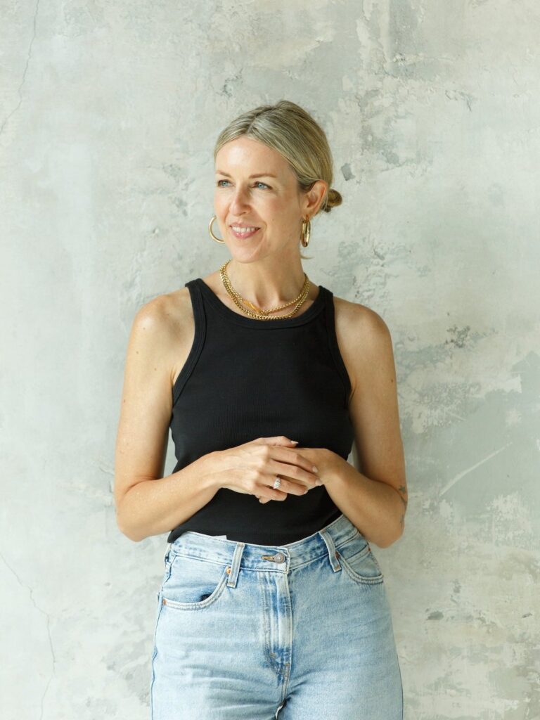 Woman with blonde hair in a low bun, wearing a black sleeveless top and light blue jeans, accessorized with gold hoop earrings and a necklace, stands against a textured light gray wall where opposites align in effortless style. - Nuovo Salon Group in Sarasota & Bradenton, FL