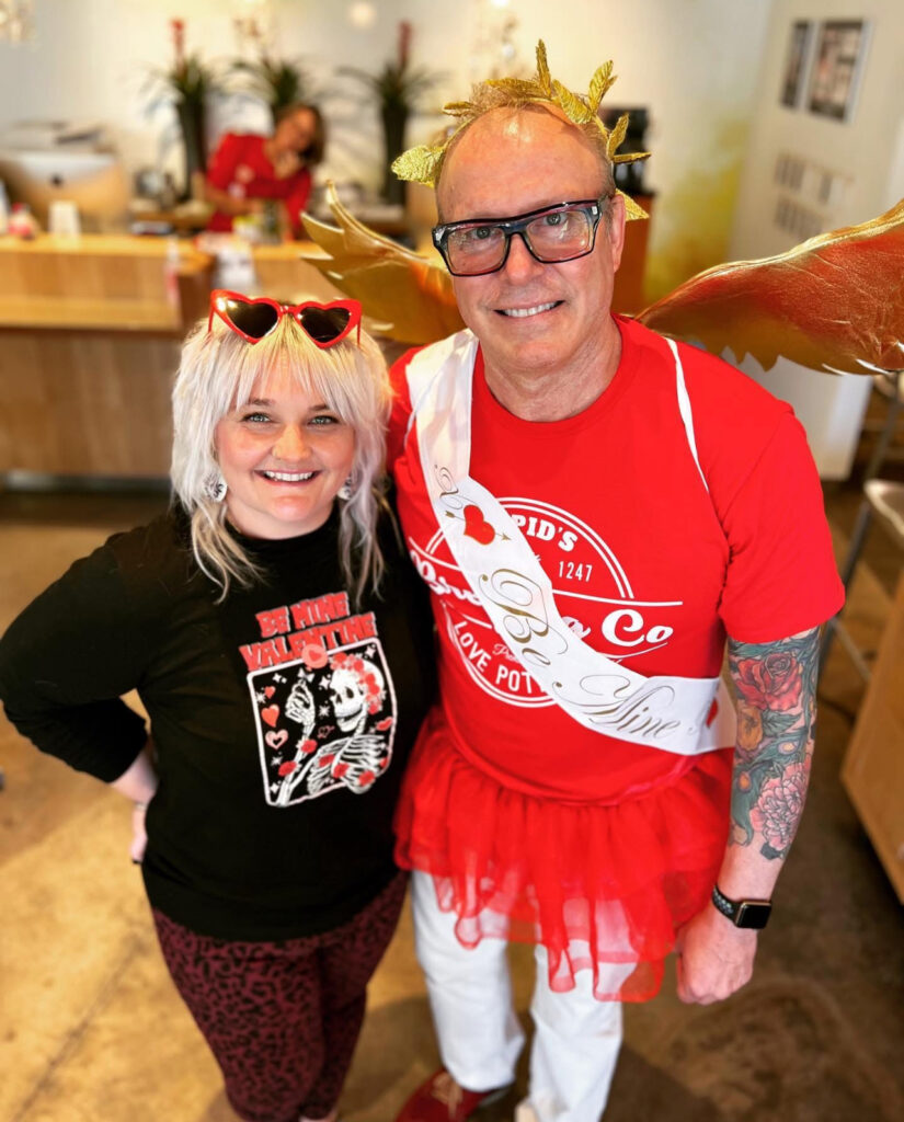 Two adults pose indoors; one wears heart-shaped glasses and a skeleton shirt, the other is dressed in red with gold wings, a sash, and a tutu, both smiling at the camera. - Nuovo Salon Group in Sarasota & Bradenton, FL