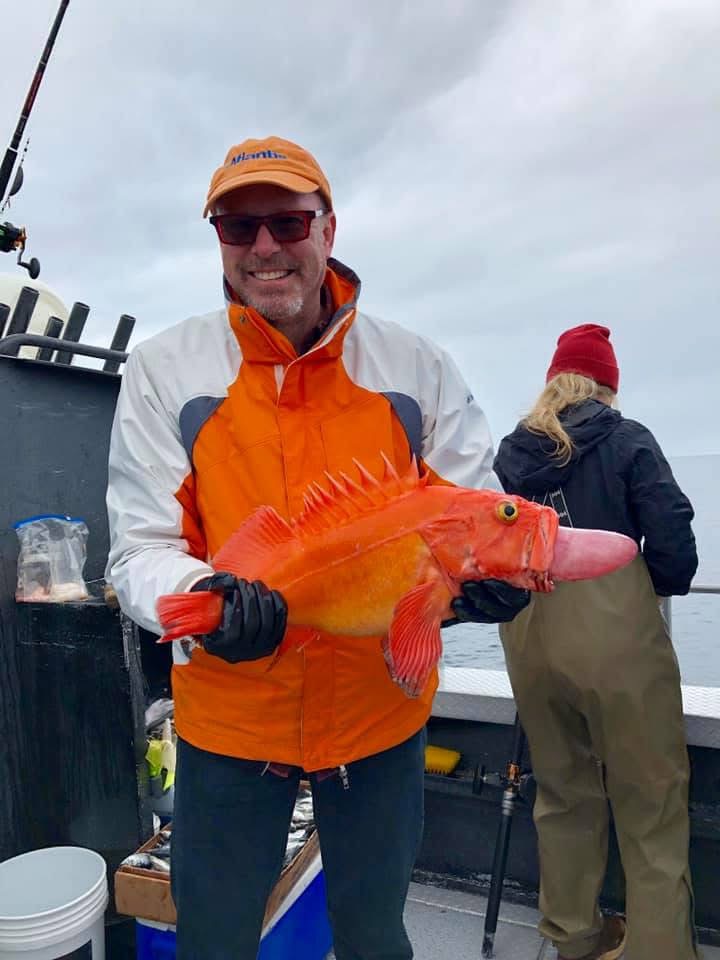 A person wearing an orange jacket and cap holds a bright orange fish with unusual features on a boat; another person stands in the background. - Nuovo Salon Group in Sarasota & Bradenton, FL