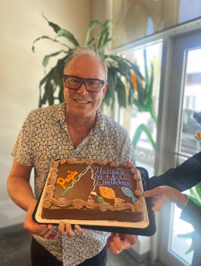 A man wearing glasses and a patterned shirt smiles while holding a chocolate birthday cake with “Happy Birthday” written in icing. - Nuovo Salon Group in Sarasota & Bradenton, FL