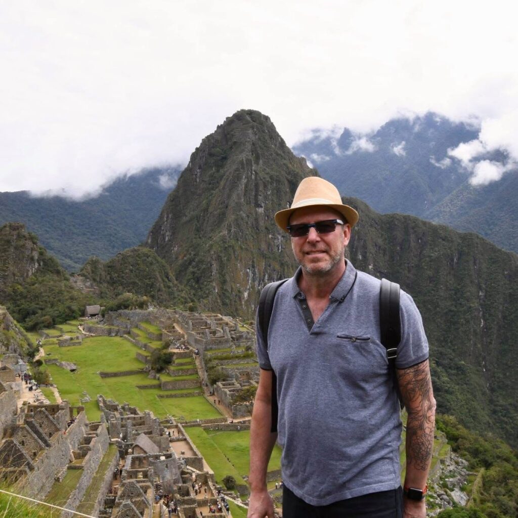 A man wearing sunglasses, a hat, and a backpack stands in front of the ancient ruins of Machu Picchu with mountains and clouds in the background. - Nuovo Salon Group in Sarasota & Bradenton, FL