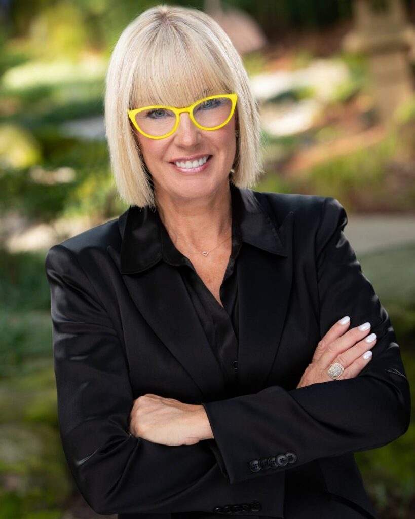 A woman with short blonde hair and yellow glasses wears a black blazer, stands outdoors with arms crossed, and smiles at the camera—proof that sometimes opposites align in perfect harmony. - Nuovo Salon Group in Sarasota & Bradenton, FL