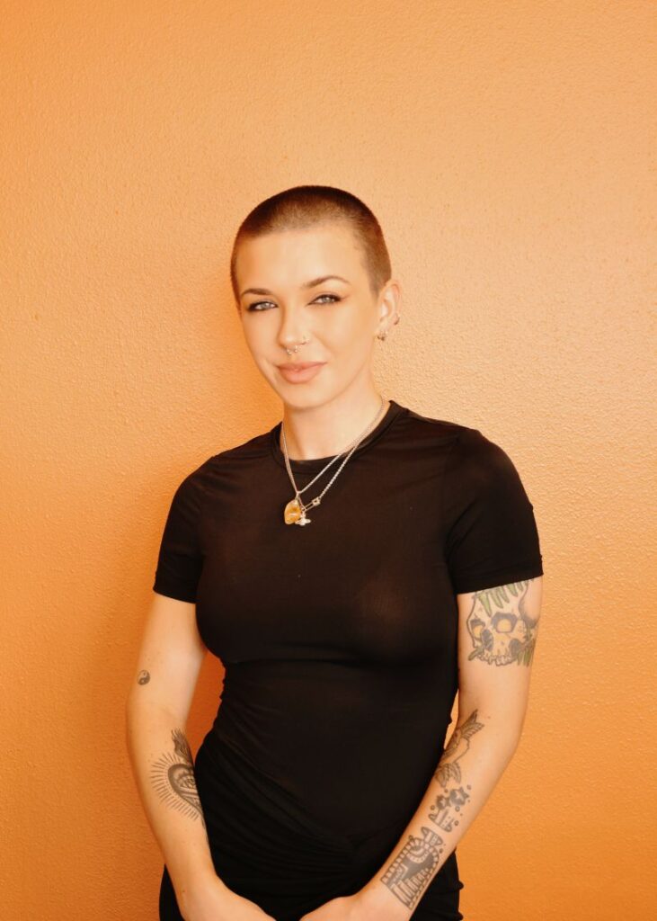 Person with a buzz cut and visible tattoos stands against an orange wall, wearing a fitted black t-shirt and layered necklaces—proof that when opposites align, bold style statements are made. - Nuovo Salon Group in Sarasota & Bradenton, FL