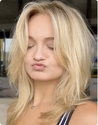 A person with layered blonde hair and closed eyes poses with puckered lips, wearing a striped top and a necklace, standing in front of a glass building. - Nuovo Salon Group in Sarasota & Bradenton, FL