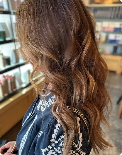 A woman with long, wavy light brown hair is seen from the side, wearing a dark embroidered top, standing indoors near shelves with bottles. - Nuovo Salon Group in Sarasota & Bradenton, FL
