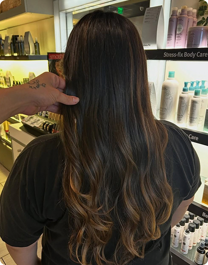 Person with long, wavy brown hair seen from behind in a store, with a hand holding a section of their hair near illuminated shelves of beauty products. - Nuovo Salon Group in Sarasota & Bradenton, FL