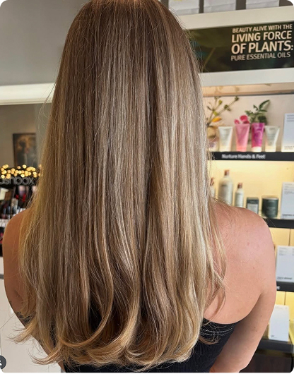 Person with long, straight, blonde hair seen from the back, standing in a salon with shelves of hair and skincare products in the background. - Nuovo Salon Group in Sarasota & Bradenton, FL