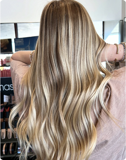 Person with long, wavy, blonde hair facing away from the camera, wearing a light pink top, in a well-lit indoor setting with cosmetic products visible in the background. - Nuovo Salon Group in Sarasota & Bradenton, FL
