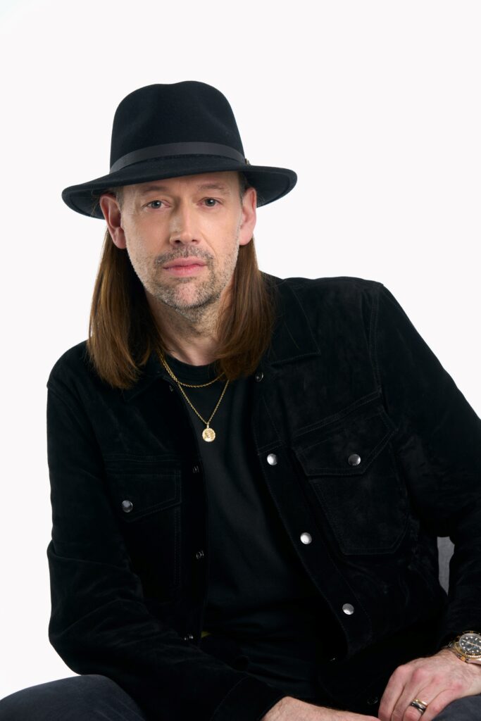 A man with long brown hair wearing a black hat, black jacket, black shirt, and gold necklaces sits against a plain white background, where opposites align in striking harmony. - Nuovo Salon Group in Sarasota & Bradenton, FL