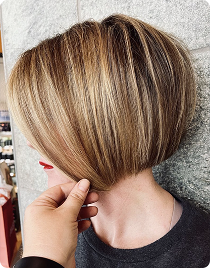 A person with straight, short, blonde hair styled in a sleek angled bob; another hand is holding a section to show the cut’s shape against a stone wall background. - Nuovo Salon Group in Sarasota & Bradenton, FL