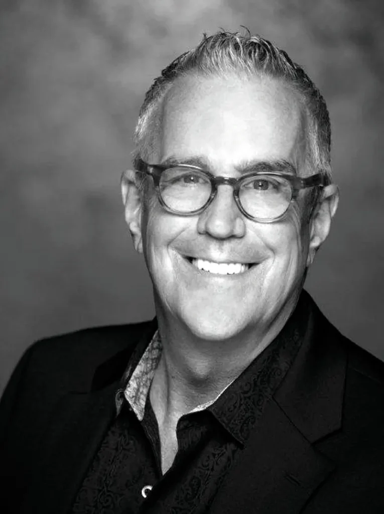 A middle-aged man with short gray hair and glasses smiles at the camera, wearing a dark jacket and patterned shirt. This black-and-white photo captures how opposites align in his classic yet modern style. - Nuovo Salon Group in Sarasota & Bradenton, FL