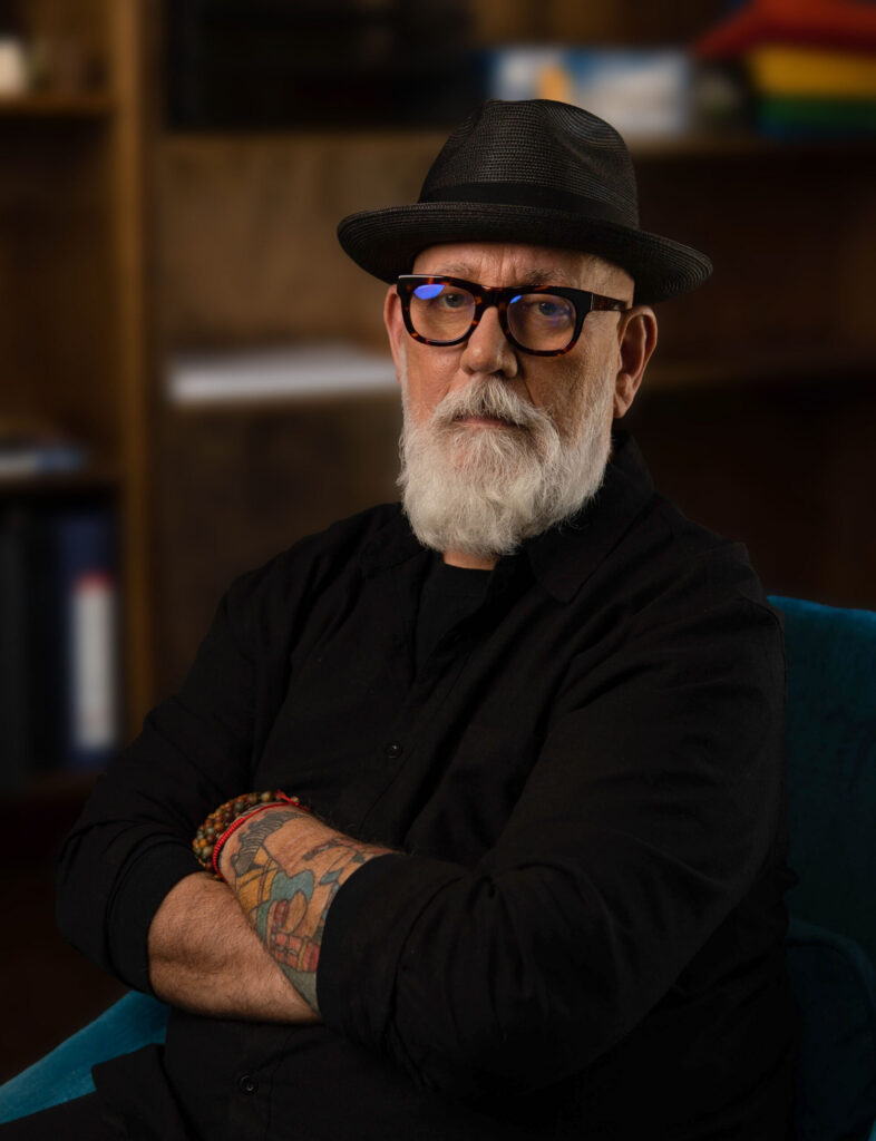 An older man with a white beard and glasses, wearing a black shirt and hat, sits with arms crossed, displaying tattoos on his forearm. Shelves with books and decor in the background hint that opposites align in his unique style. - Nuovo Salon Group in Sarasota & Bradenton, FL