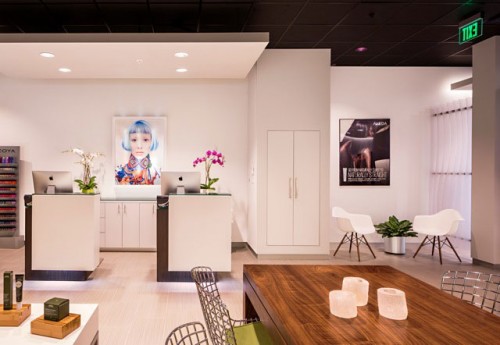 Modern reception area with white desks, two iMacs, orchid plants, wall art, and chairs. A large wooden table with candles sits in the foreground. - Nuovo Salon Group in Sarasota & Bradenton, FL