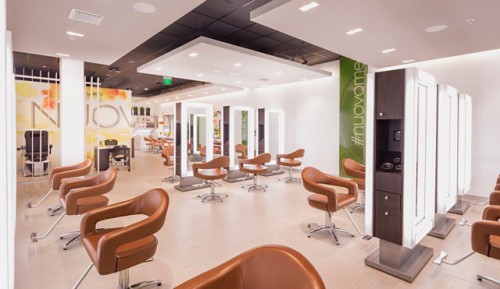 A modern hair salon with brown styling chairs, large mirrors, and bright lighting; the space is clean, open, and features a green accent wall with the word "#nuovome. - Nuovo Salon Group in Sarasota & Bradenton, FL