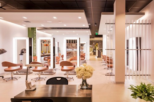 Modern hair salon interior with multiple styling stations, brown chairs, mirrors, and bright lighting. A desk with flowers and a plant is visible in the foreground. - Nuovo Salon Group in Sarasota & Bradenton, FL