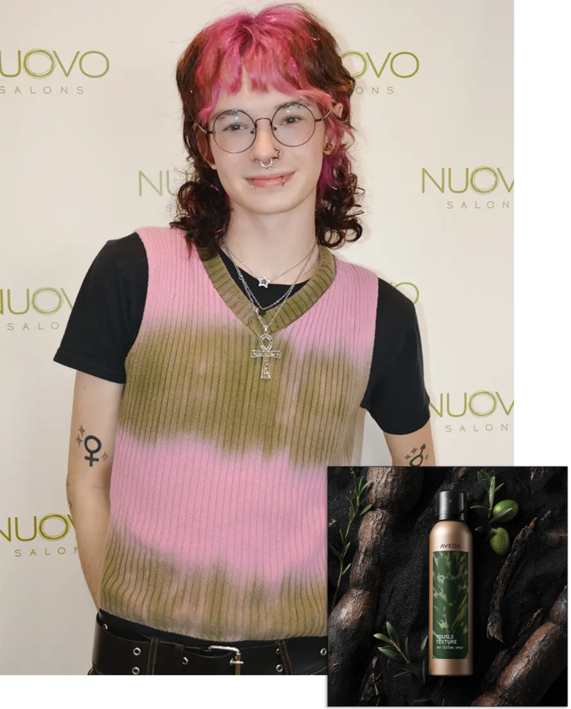Person with short, curly pink and brown hair, glasses, and layered necklaces stands in front of a Nuovo Salons backdrop; inset shows a bottle of Aveda styling product. - Nuovo Salon Group in Sarasota & Bradenton, FL