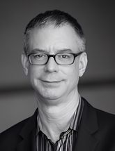 A man with short hair and glasses, wearing a striped shirt and a blazer, looks at the camera with a neutral expression in this black and white photo—where opposites align in timeless style. - Nuovo Salon Group in Sarasota & Bradenton, FL
