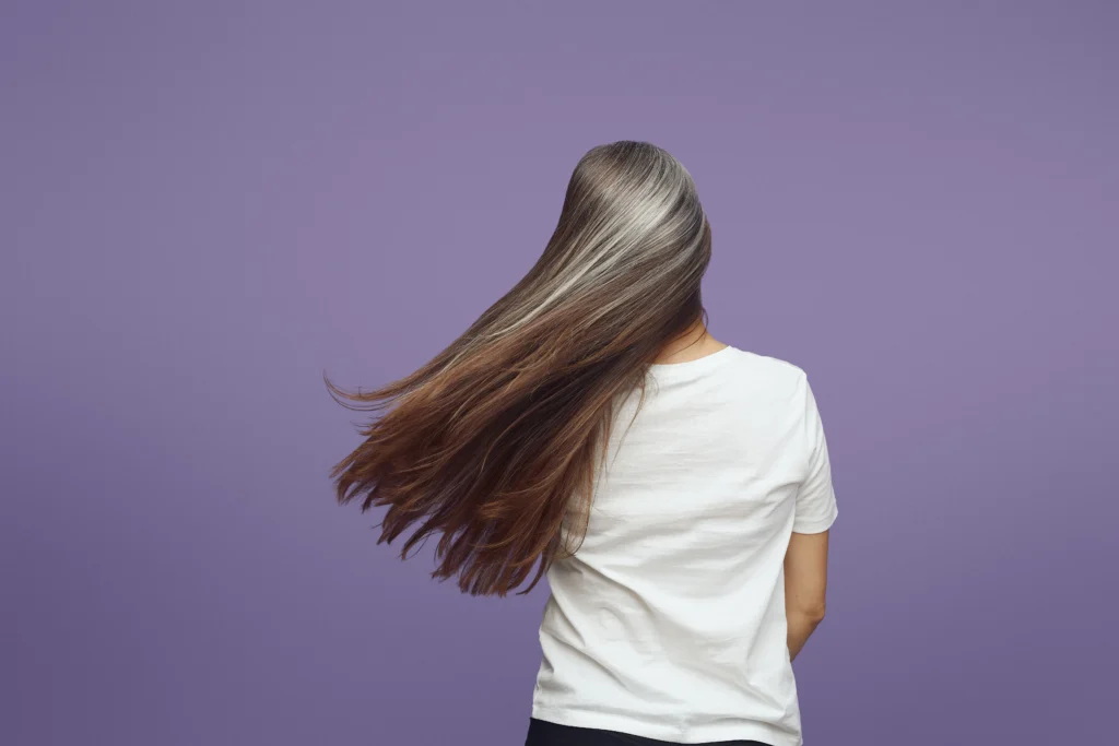 A person seen from behind with long, flowing brown and grey hair, wearing a plain white t-shirt against a purple background. - Nuovo Salon Group in Sarasota & Bradenton, FL